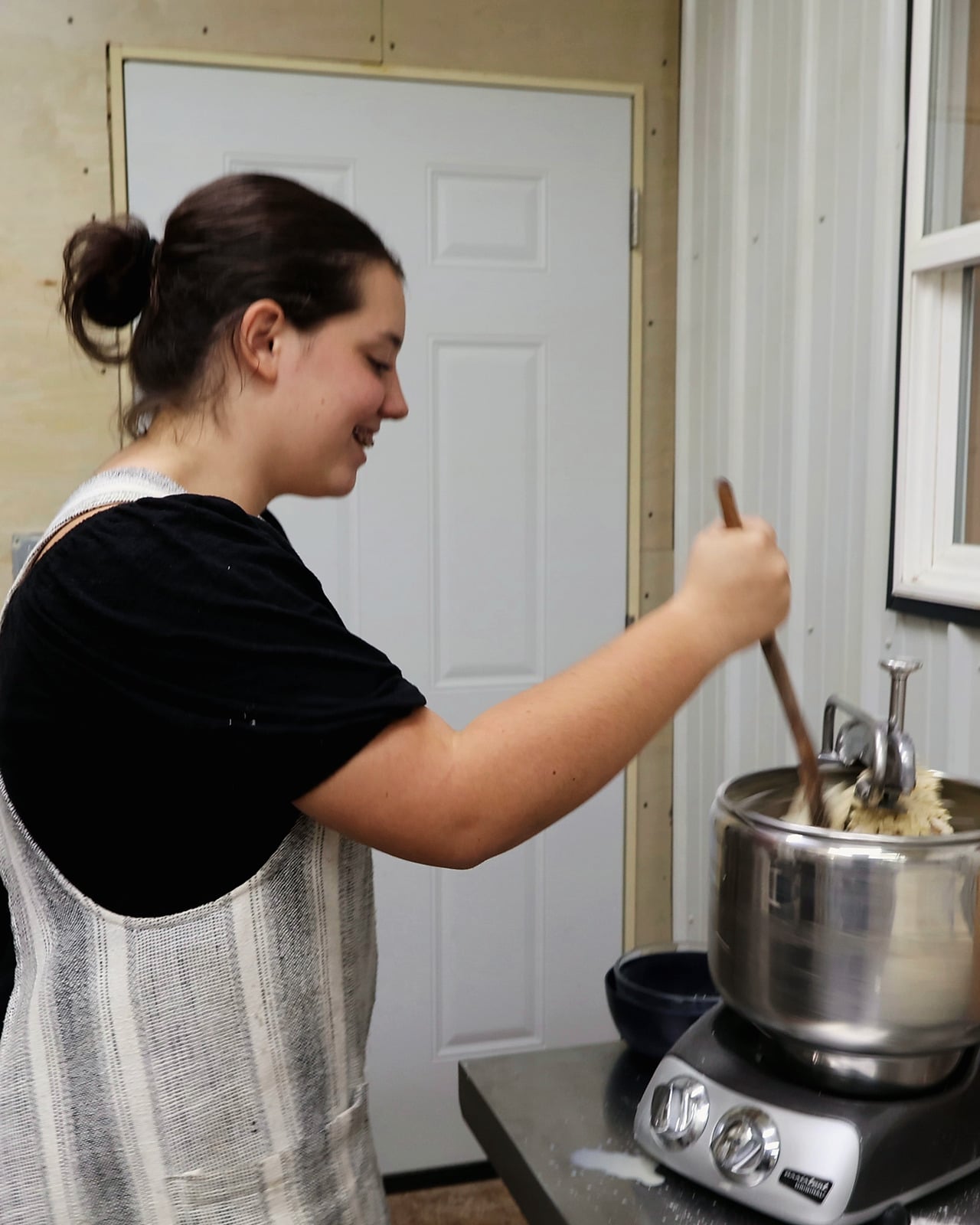 Avery, in a black top and a grey-and-cream striped linen apron with her hair in a low bun, smiles as she looks down and stirs cinnamon-roll dough in a steel stand mixer with a long wooden spoon, with a white door and a window behind her.