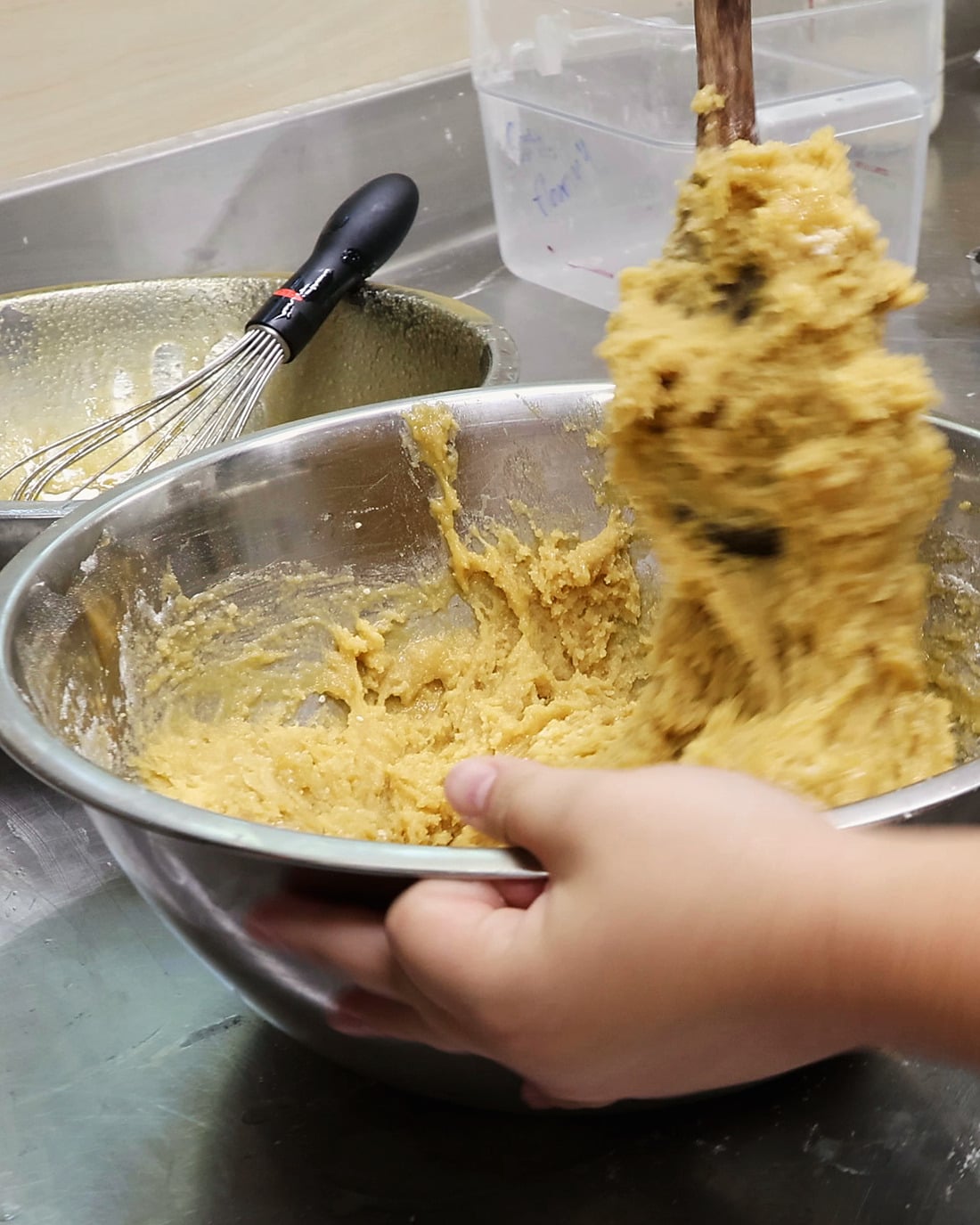 A dark wooden spoon lifts a thick fold of golden chocolate-chip cookie dough out of a steel mixing bowl. A hand tilts the bowl toward the camera, and a second bowl with a whisk sits behind it.