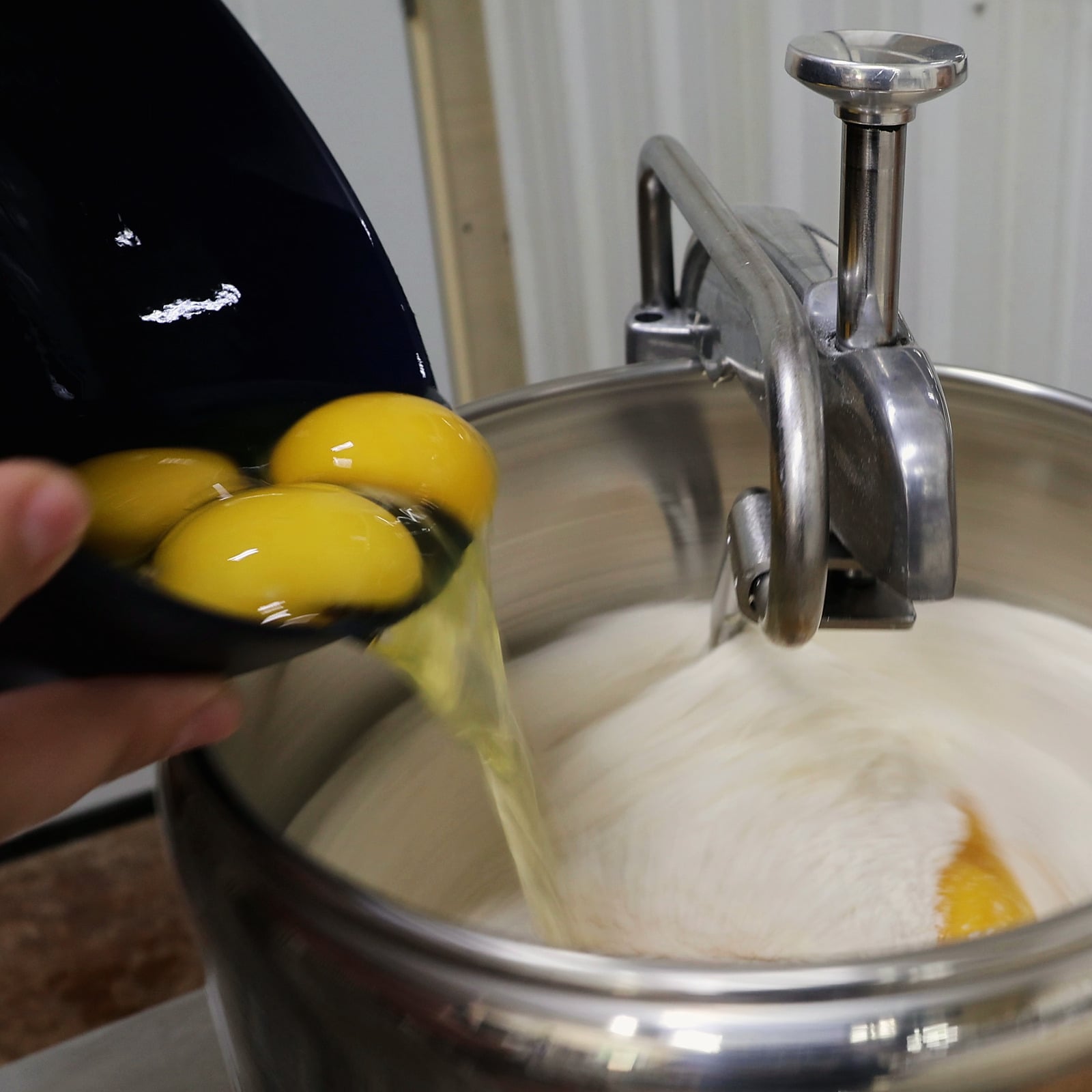 Golden egg yolks slide from a tipped navy bowl into white flour turning in a stainless-steel mixer bowl, trailing a glossy strand of egg white; one yolk has already landed in the flour.