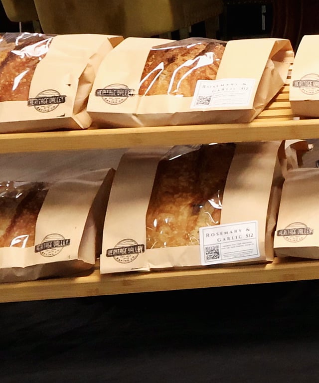 Two Heritage Valley kraft window bags labelled Rosemary & Garlic sit one above the other on a bamboo market shelf, with golden, blistered sourdough crusts showing through the cellophane. Bagged plain sourdough loaves sit on either side and a black tablecloth runs below.