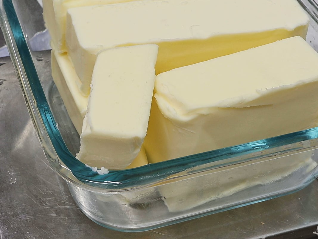 Close-up of pale, creamy sticks of butter stacked in a square glass dish with a teal-tinted rim.