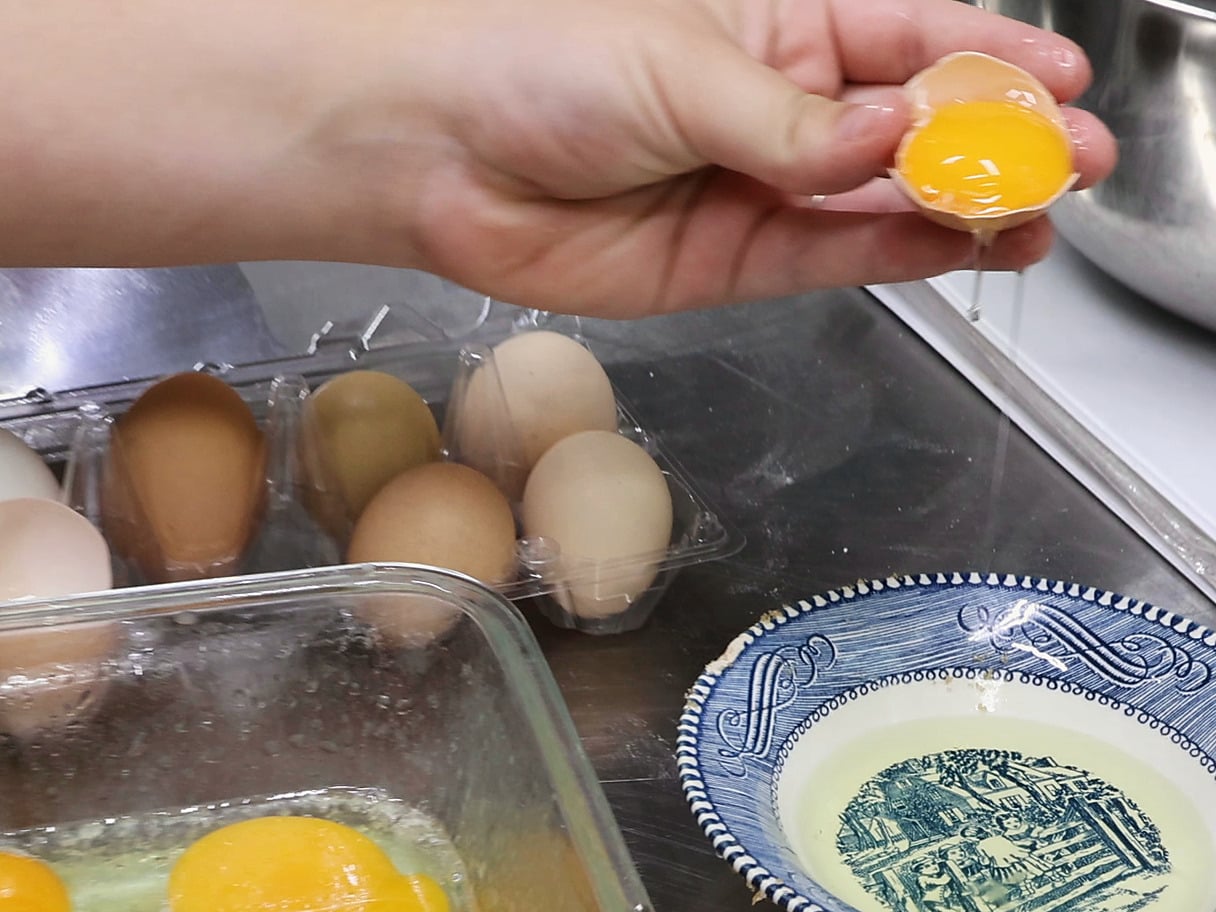 A hand holds a cracked brown eggshell with a bright golden yolk above a carton of brown, cream and olive farm eggs. A thread of white drips into a blue-and-white transferware bowl, and a glass dish of separated yolks sits in the foreground.