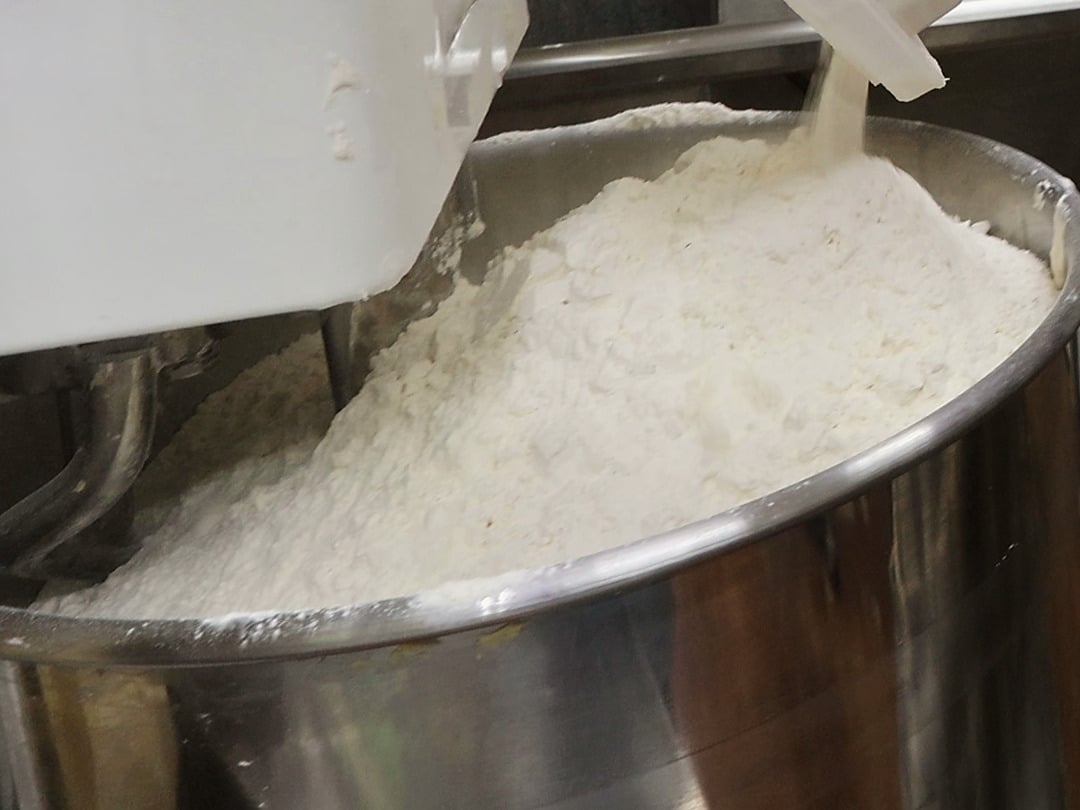 A heaped mound of fresh-milled flour in the steel bowl of a spiral mixer. A thin stream of flour pours from the lip of a white tub onto the peak, with the dough hook and mixer head at the left.
