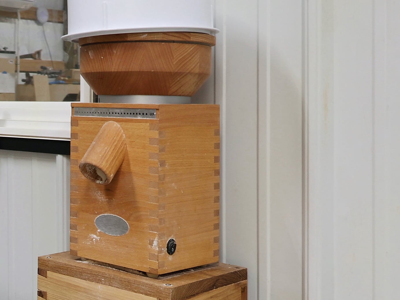 A wooden grain mill with dovetailed beech corners, a turned wooden crank handle and a wooden hopper bowl under a white hopper extension. It stands on a wooden block against white panelled walls in the bakehouse, next to a window.