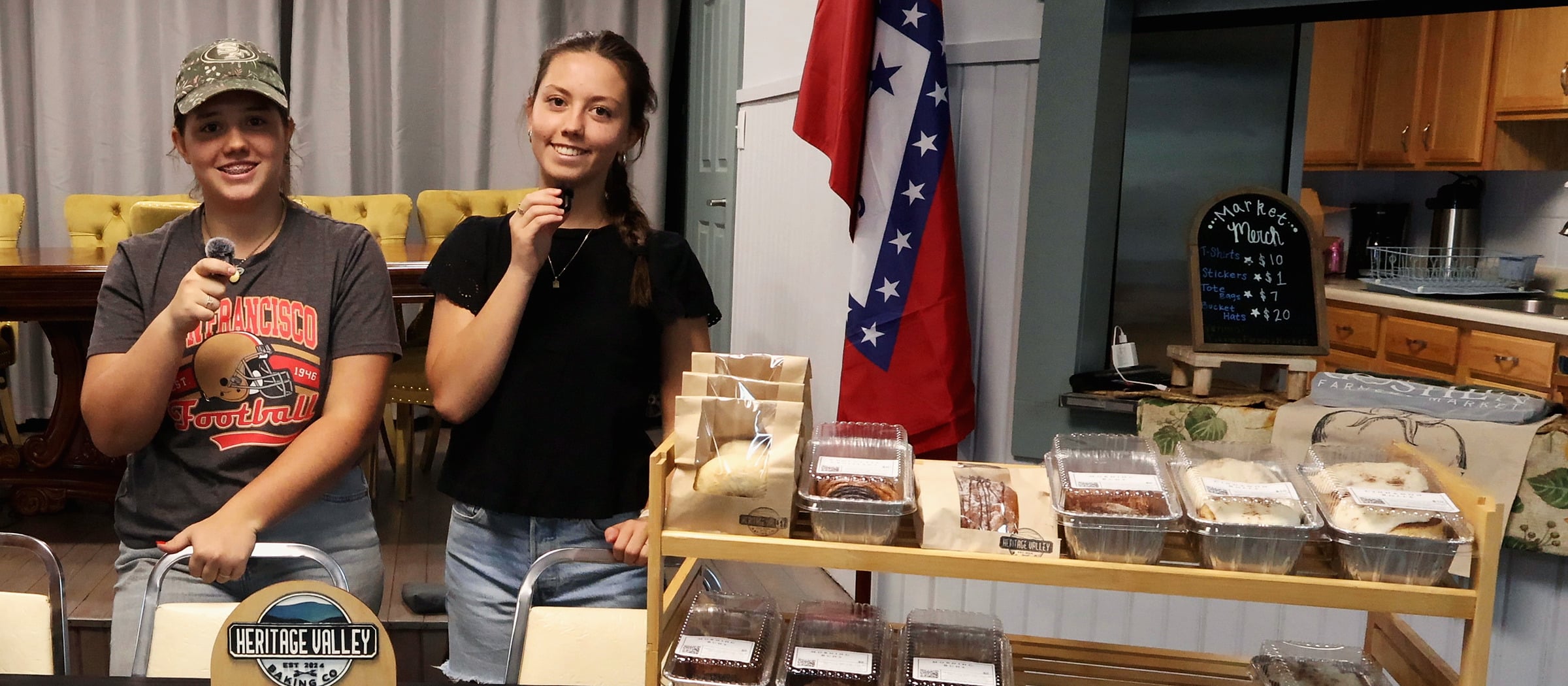 Olivia and Avery smile at the camera from behind their Heritage Valley Baking Co. market table. Beside them a bamboo shelf holds a cinnamon-swirl loaf, bagged rolls and trays of iced cinnamon rolls, and the arched wooden Heritage Valley sign stands at the front of the table.