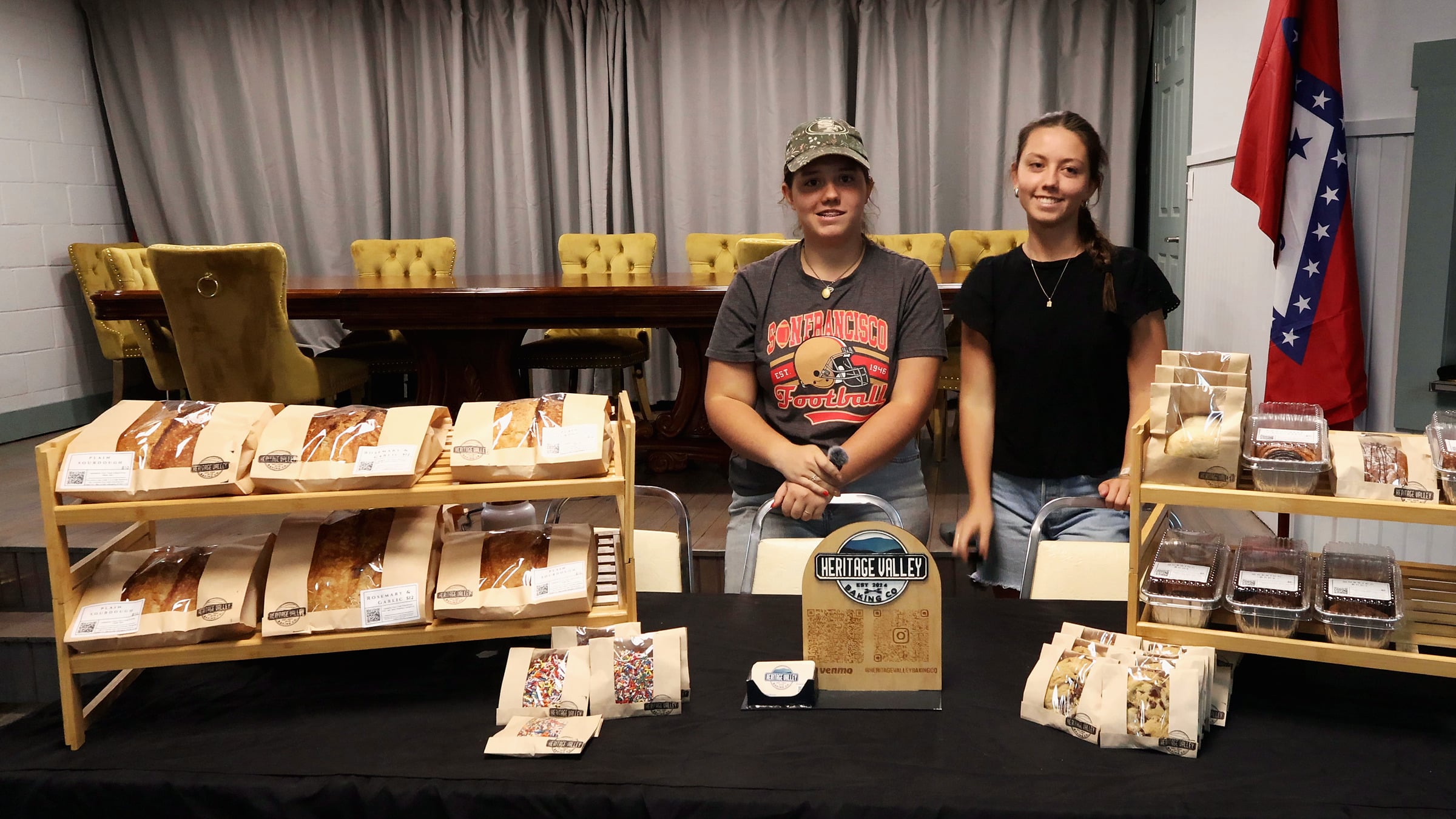 Sisters Avery and Olivia stand behind the Heritage Valley Baking Co. market table and smile at the camera. On the left, a bamboo shelf holds kraft window bags of Plain Sourdough and Rosemary and Garlic loaves. In the centre is the arched wooden Heritage Valley sign, with bags of sprinkle cookies in front. On the right, a second shelf holds cinnamon rolls, a cinnamon-swirl loaf and quick breads in clear-lid pans, with chocolate-chip cookie bags on the black tablecloth. Grey curtains, gold tufted chairs and an Arkansas flag fill the background.