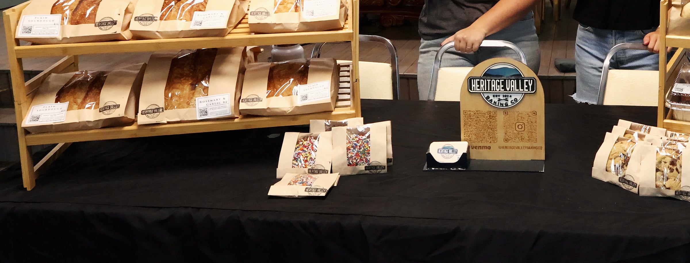 The Heritage Valley Baking Co. market table on a black tablecloth. On the left, a bamboo shelf holds kraft window bags of Plain Sourdough and Rosemary and Garlic loaves. In the middle are bags of sprinkle cookies, a card holder and the wooden Heritage Valley sign. Chocolate-chip cookie bags sit at the right. The two sisters stand behind the table, shown from the waist down.
