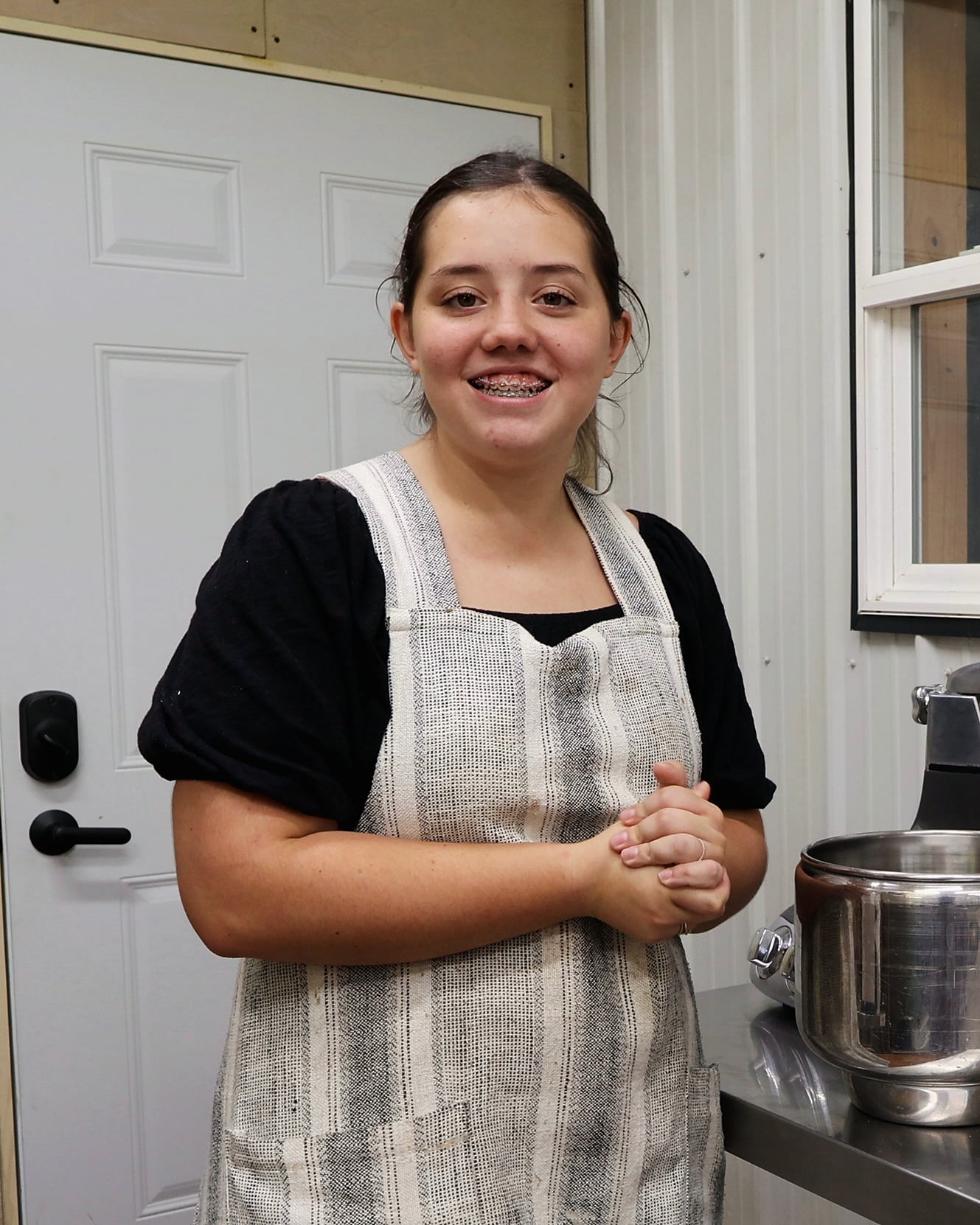 Avery smiling at the camera in the bakehouse kitchen. She wears a black puff-sleeve top under a grey-and-cream striped linen apron, with her hands clasped and a steel mixer bowl beside her.
