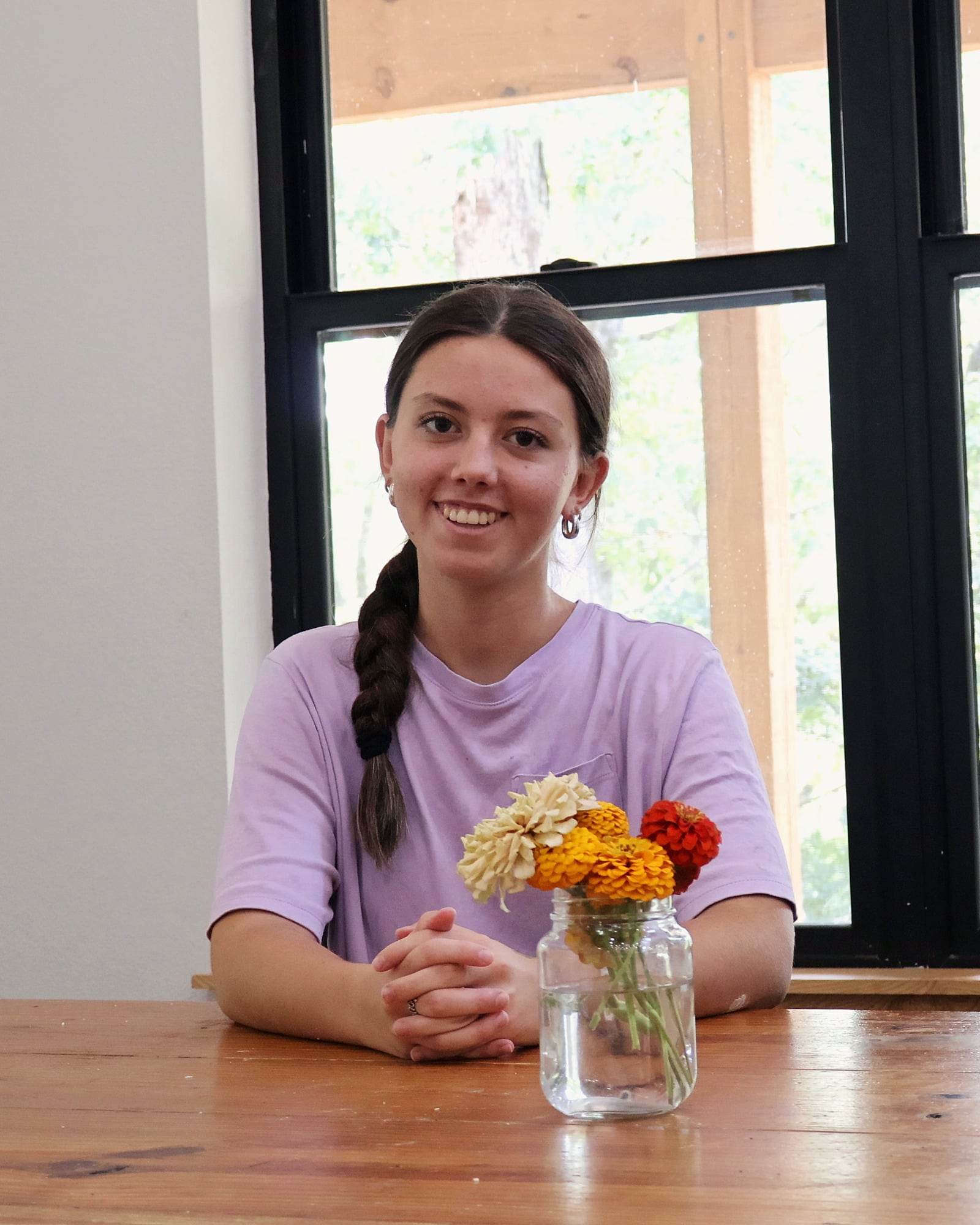 Olivia, in a lilac tee with her long brown braid over one shoulder and silver hoop earrings, smiles at the camera. She sits at a honey-pine table with her hands clasped beside a glass jar of zinnias, in front of a black-framed window with trees outside.